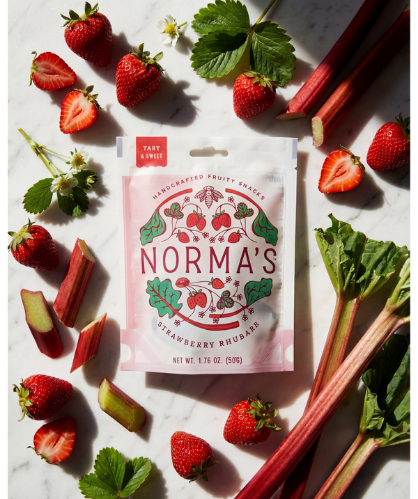 Norma's strawberry rhubarb fruit snack package surrounded by strawberries and rhubarb on a marble surface.